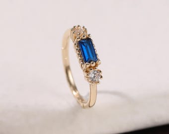 Könnte beinhalten: Ein goldener Ring mit einem rechteckigen, tiefblauen Edelstein als Mittelpunkt, flankiert von kleineren, klaren Edelsteinen. Der Ring hat ein zartes Band und ist vor einem neutralen Hintergrund platziert.