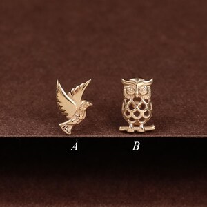 May include: Two gold-coloured pins: a flying bird and an owl. The bird pin has outstretched wings, while the owl pin has a detailed, openwork design. Both pins are displayed on a dark brown surface.