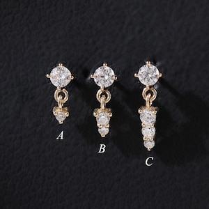 以下が含まれることがあります： クリアで輝く宝石が付いた、3つのゴールドカラーのイヤリング。各イヤリングは異なるデザインで、ぶら下がっている宝石の数が異なります。イヤリングは暗い背景に展示されており、その輝きを際立たせています。