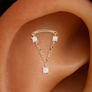 Op de afbeelding: Een goudkleurige oorbel met drie heldere, vierkant geslepen edelstenen. De bovenkant van de piercing heeft een gebogen staafje met twee edelstenen, en een ketting met een derde edelsteen hangt eronder. De piercing wordt op een oor getoond.