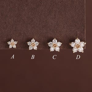 May include: Four flower-shaped stud earrings labeled A, B, C, and D in increasing size. Each earring features five clear white petals surrounding a small central amber-colored stone, set in a metallic frame against a dark brown background.