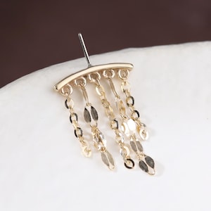 May include: A gold earring featuring a curved top and hanging chain details. The chains are decorated with small, oval charms. The earring is set against a white surface with a brown backdrop.