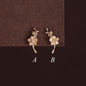 May include: Two gold-toned metal earrings shaped like small flower sprigs, labeled A and B. Each earring features a five-petaled blossom with a central detail and a curved stem, presented against a dark brown background for clear visual contrast.