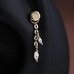 May include: A gold-colored earring with a small, round stud and a dangling chain. The chain holds three small, teardrop-shaped, clear gemstones. The earring is shown against a black background.