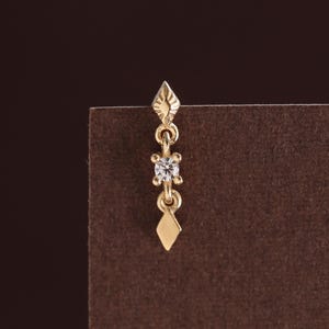 May include: A gold-toned dangle earring featuring a small, pointed diamond-shaped top element, a central clear round gemstone set in a four-prong frame, and a matching pointed diamond-shaped charm at the bottom, all connected by thin metal links.