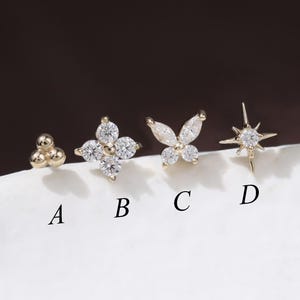 May include: Four gold-coloured stud earrings with various designs: a cluster of three small spheres, a four-petal flower, a butterfly, and a starburst. Each earring features clear, sparkling gemstones. The earrings are displayed on a white surface.