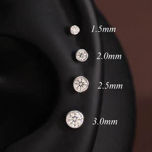 Może przedstawiać: Cztery okrągłe srebrne kolczyki z przezroczystymi kamieniami ułożone pionowo na ciemnym modelu ucha. Rozmiary są oznaczone jako 1,5 mm, 2,0 mm, 2,5 mm i 3,0 mm, wskazując średnicę każdego pojedynczego kolczyka widocznego na zdjęciu.