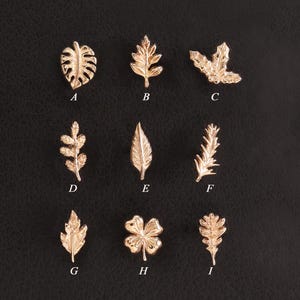 May include: A collection of nine small rose gold-toned metal leaf and plant-shaped charms arranged in a three-by-three grid. Each charm is labeled with a letter from A to I, featuring distinct botanical designs including ferns, clover, and various leaves.