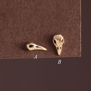 May include: Two small gold-toned metal charms shaped like bird skulls. The charm labeled A is shown in profile, while the charm labeled B is shown from a front-facing perspective. Both pieces feature hollow eye sockets and a pointed beak.