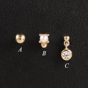 Könnte beinhalten: Drei goldfarbene Ohrringe auf schwarzem Untergrund. Sie umfassen einen kleinen Kugelstecker, einen Stecker mit perlenartigem Mittelteil und einen hängenden Ohrring mit einem klaren, runden Stein. Die Ohrringe sind mit A, B und C beschriftet.