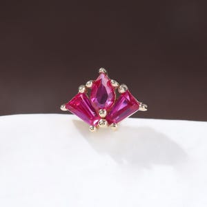 May include: A small, gold-toned jewelry piece featuring a cluster of vibrant pink gemstones. The design includes a central, teardrop-shaped stone flanked by rectangular gems, creating a symmetrical, floral-like arrangement. The piece is set against a white surface.