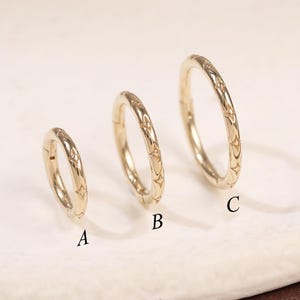 May include: Three gold-colored hoop earrings of varying sizes, labeled A, B, and C. Each earring features a repeating star pattern. The earrings are displayed on a white, oval-shaped surface.