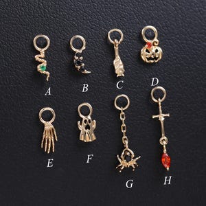Pode incluir: Uma coleção de amuletos dourados com tema de Halloween. Os amuletos incluem uma cobra, uma aranha, uma abóbora, uma mão, um fantasma, uma aranha, uma cruz e uma mão. Os amuletos são exibidos em uma superfície preta.