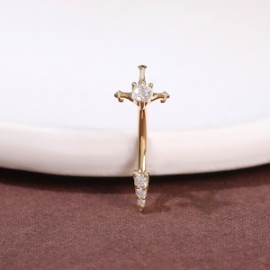 Peut inclure: Un bijou de corps en forme d'haltère courbé en or avec un motif d'épée. La garde est ornée d'une pierre précieuse ronde transparente, tandis que la lame se termine en pointe avec trois petites pierres transparentes alignées verticalement.