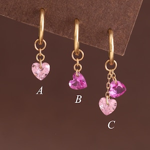 Pode incluir: Três brincos de argola dourados com pingentes em forma de coração. O brinco A tem um coração rosa claro, o B um coração rosa escuro e o C apresenta dois corações pendentes em rosa claro e escuro. Cada brinco é marcado com uma letra.