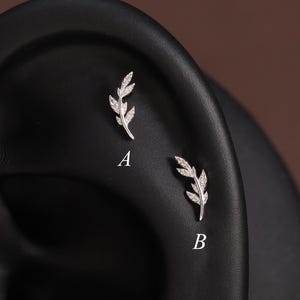 May include: Two silver-toned leaf-shaped ear studs, labelled A and B, featuring small clear stones set along the branches. The jewellery is displayed on a black ear model, with design A positioned higher than design B, showcasing a delicate botanical aesthetic.