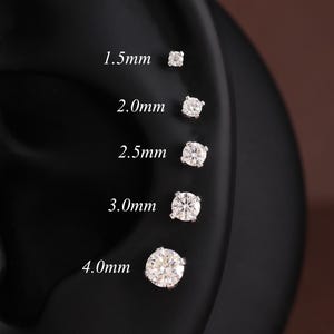 May include: Five round, clear stud earrings arranged vertically on a black ear model. The sizes are labeled as 1.5mm, 2.0mm, 2.5mm, 3.0mm, and 4.0mm. Each earring features a single faceted stone set in a metallic prong setting, showcasing a range of diameters.