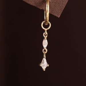 May include: A single gold earring with a dangling design. The earring features a small hoop at the top, connected to a chain with two small, elongated, clear stones and a larger, diamond-shaped clear stone at the bottom. The earring is set against a brown background.