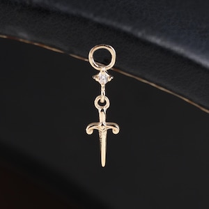 May include: A gold-colored earring with a small sword pendant. The earring features a small diamond-like accent and a circular top for attachment. The sword pendant has a detailed hilt and blade, creating a delicate and elegant design.