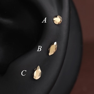 May include: Three small gold-toned stud earrings with faceted surfaces are displayed on a black ear model. The earrings are labeled with white letters A, B, and C, showing a round shape, a marquise shape, and a teardrop shape respectively.