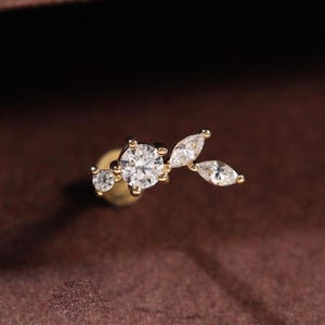 Peut inclure: Une boucle d'oreille à tige en or ornée d'un groupe de pierres transparentes. Le design comprend une grande pierre ronde taille brillant, une plus petite pierre ronde et deux pierres taille marquise disposées en forme de branche.