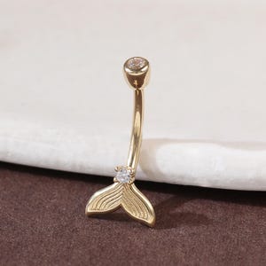 Puede incluir: Un piercing para el ombligo de color dorado con un diseño de cola de sirena. La barra curva presenta una pequeña gema transparente en la parte superior y una gema transparente más grande en la cola. La aleta caudal está detallada con líneas curvas.