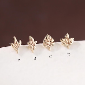 May include: Four gold-colored leaf-shaped stud earrings, labeled A, B, C, and D, are displayed on a white surface. Each earring features a different leaf design, showcasing intricate details. The background is a gradient of brown.