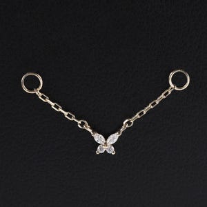 Puede incluir: Una delicada joya corporal dorada con un colgante de mariposa con piedras preciosas transparentes. La pieza tiene dos extremos circulares conectados por una cadena, creando un diseño simétrico. La joya está sobre un fondo oscuro.