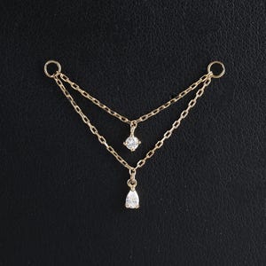 Puede incluir: Una delicada cadena de cuerpo dorada con dos hilos, cada uno adornado con una gema brillante. El hilo superior presenta una gema redonda, mientras que el hilo inferior muestra una gema en forma de lágrima. La cadena está diseñada para ser usada sobre el cuerpo.