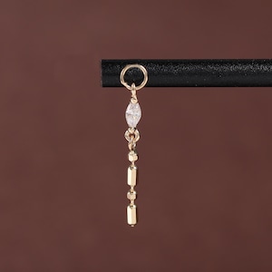 Pode incluir: Um brinco dourado com um design pendurado. O brinco apresenta uma pequena pedra preciosa transparente em forma de marquesa no topo, seguida por uma série de pequenas barras retangulares douradas. O brinco é suspenso por um pequeno círculo.