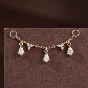 May include: A gold chain connector piece featuring two circular end loops, three dangling pear-shaped clear stones, and two small clusters of three tiny clear stones set along the chain, creating a delicate and symmetrical jewellery component.