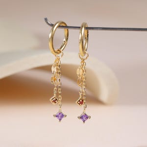 Pode incluir: Brincos de argola dourados com correntes penduradas. Cada brinco apresenta pequenos discos dourados, uma gema quadrada vermelha e uma gema quadrada roxa. Exibidos em um fundo neutro.