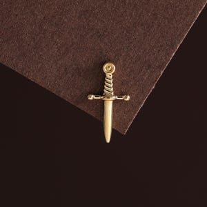 May include: A gold-toned metal charm shaped like a small sword or dagger. The piece features a twisted handle design, a horizontal crossguard with rounded ends, and a smooth, pointed blade. The charm includes a small circular loop at the top.