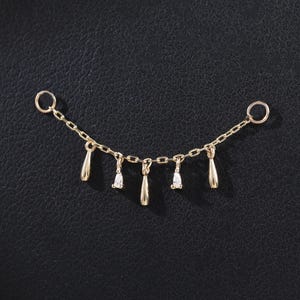Puede incluir: Una cadena dorada con colgantes en forma de lágrima y diamante. La cadena tiene un pequeño anillo en cada extremo y está colocada sobre un fondo negro. Los colgantes están espaciados uniformemente.