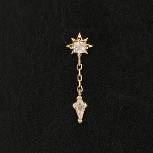 May include: A gold-toned earring featuring a starburst-shaped stud with a central square-cut clear stone. A delicate gold chain hangs from the stud, ending in a marquise-cut clear stone pendant. The design is symmetrical and metallic in finish.