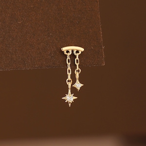 May include: A gold stud earring featuring a curved bar top with two dangling gold chains of different lengths. Each chain ends in a small, four-pointed star charm, with the longer chain featuring a clear, sparkling stone at the centre of the star.