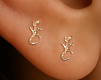 May include: A pair of gold-colored lizard stud earrings. The earrings are small and detailed, with the lizard's body and legs clearly defined. The earrings are worn on an ear, suggesting they are for pierced ears.