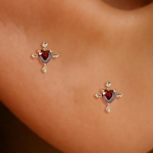 Op de afbeelding: Een paar kleine gouden oorbellen, elk met een hartvormige rode edelsteen in het midden. De oorbellen hebben een kruisvormig ontwerp met kleine, heldere accentstenen. De oorbellen worden op een oor getoond.