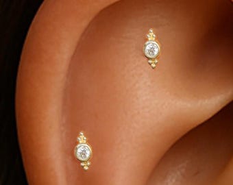 Könnte beinhalten: Nahaufnahme eines Ohrs mit zwei kleinen, goldfarbenen Ohrringen. Jeder Ohrring hat einen zentralen, klaren Edelstein, der von kleineren, dekorativen Perlen umgeben ist. Die Ohrringe sind auf der Haut des Ohrs angebracht.