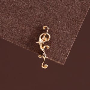 May include: A gold-toned metal earring featuring an elongated, vertical design with a central four-pointed star element, curved scrollwork, and a small circular loop at the top, showcasing a polished, reflective surface finish throughout.