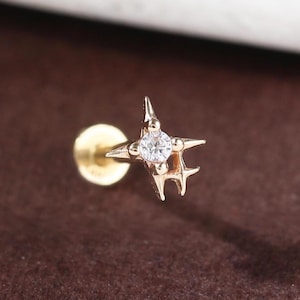 May include: A gold-colored star-shaped earring with a small, clear gemstone in the center. The earring has a flat back and a post for piercing. The star design has four points and a small, round base.
