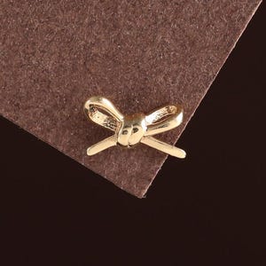May include: A small, gold-coloured bow-shaped charm. The charm is positioned on the corner of a brown, textured surface against a dark brown background. The bow has a detailed design.