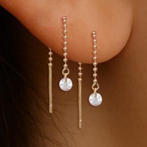May include: Gold earrings with a minimalist design. One earring has a straight bar, the other a chain of small beads. Both have a small, round, clear gemstone at the end. The earrings create an elegant and understated look.