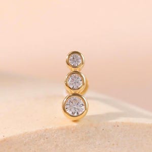 Puede incluir: Un pendiente de oro con tres piedras redondas, transparentes y facetadas dispuestas en línea vertical. Cada piedra está engastada en un bisel de oro pulido, y las piedras disminuyen ligeramente de tamaño desde la parte inferior a la superior.