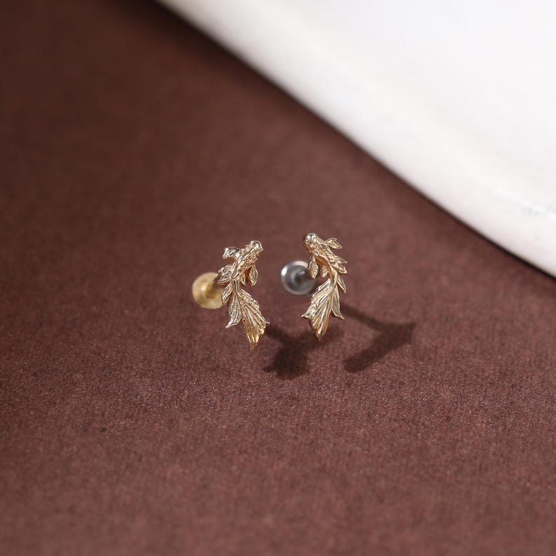 May include: A pair of gold-toned stud earrings shaped like koi fish with detailed scales and flowing fins. Each earring features a post back mechanism, with one showing a gold-colored backing and the other showing a silver-colored backing.