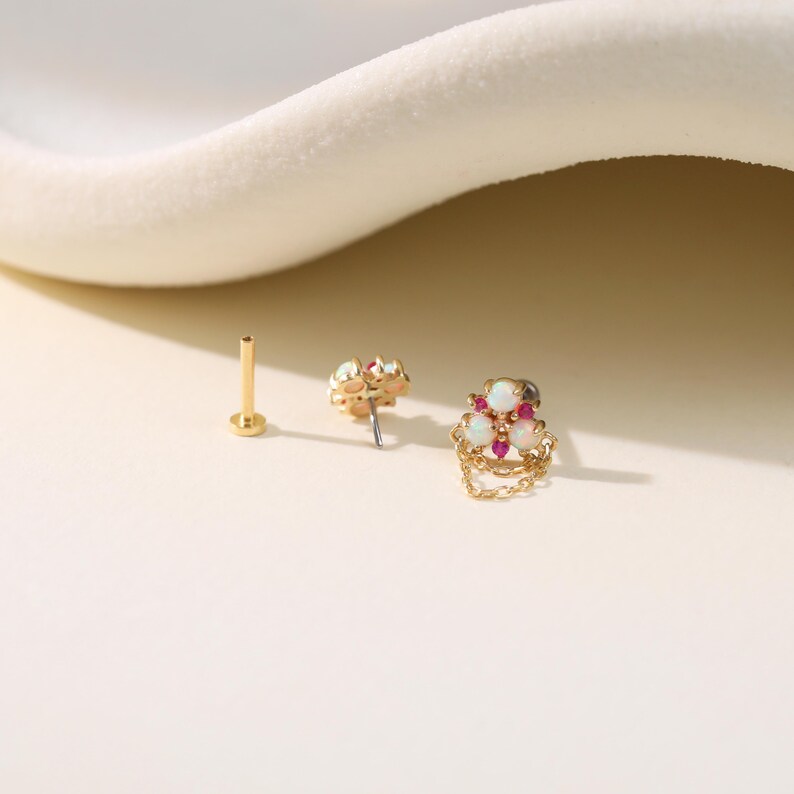 May include: A collection of gold-colored body jewelry, including a post and two decorative pieces. One piece features a cluster of white and pink gemstones with a delicate chain. The other has a similar gemstone cluster.