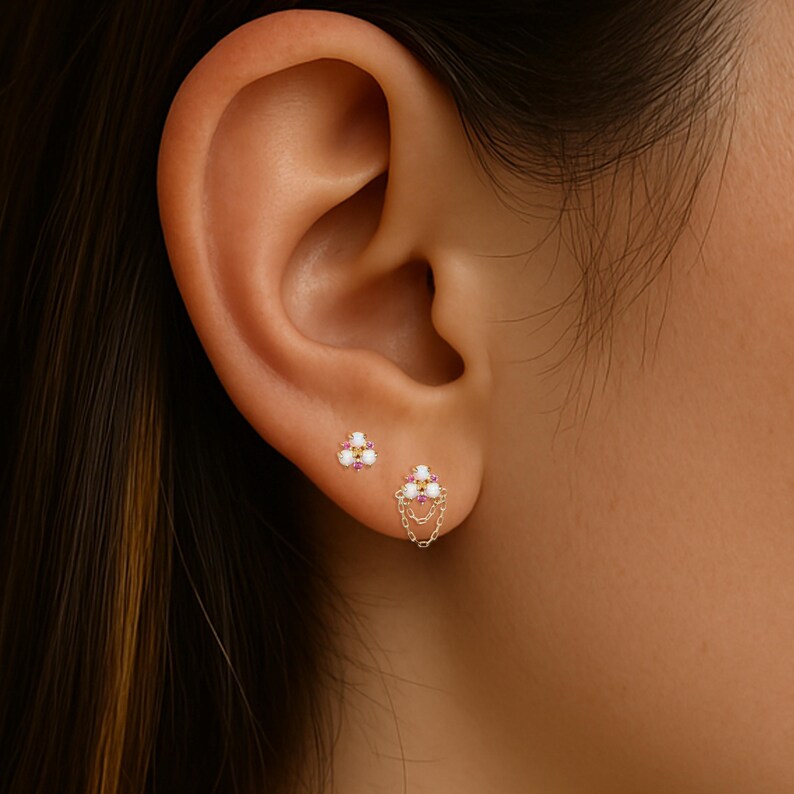 May include: Close-up of two gold earrings. The first is a small, triangular stud with white and pink stones. The second earring is a cluster of white and pink stones with a dangling chain. The earrings are set against the backdrop of an ear.