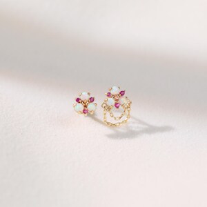 May include: Two gold earrings with white opal and small pink gemstone accents. One earring features a chain design. The earrings are set against a white background, showcasing their delicate details and craftsmanship.