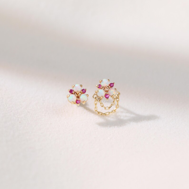 May include: Two gold earrings with white opal and small pink gemstone accents. One earring features a chain design. The earrings are set against a white background, showcasing their delicate details and craftsmanship.