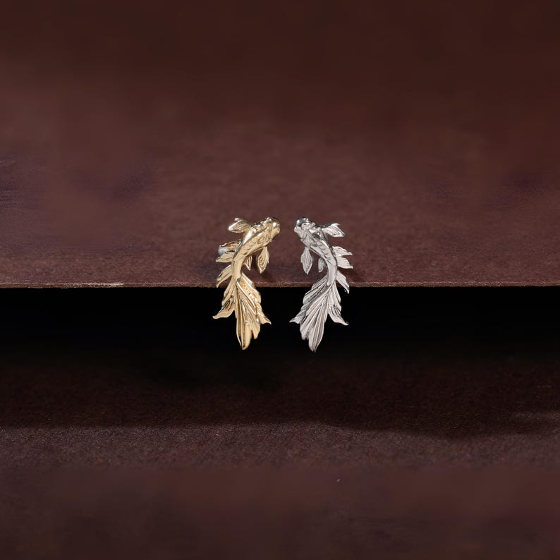 May include: A pair of stud earrings shaped like koi fish, with one earring finished in a polished gold tone and the other earring finished in a polished silver tone, featuring detailed fins and scales on the surface of each fish figure.